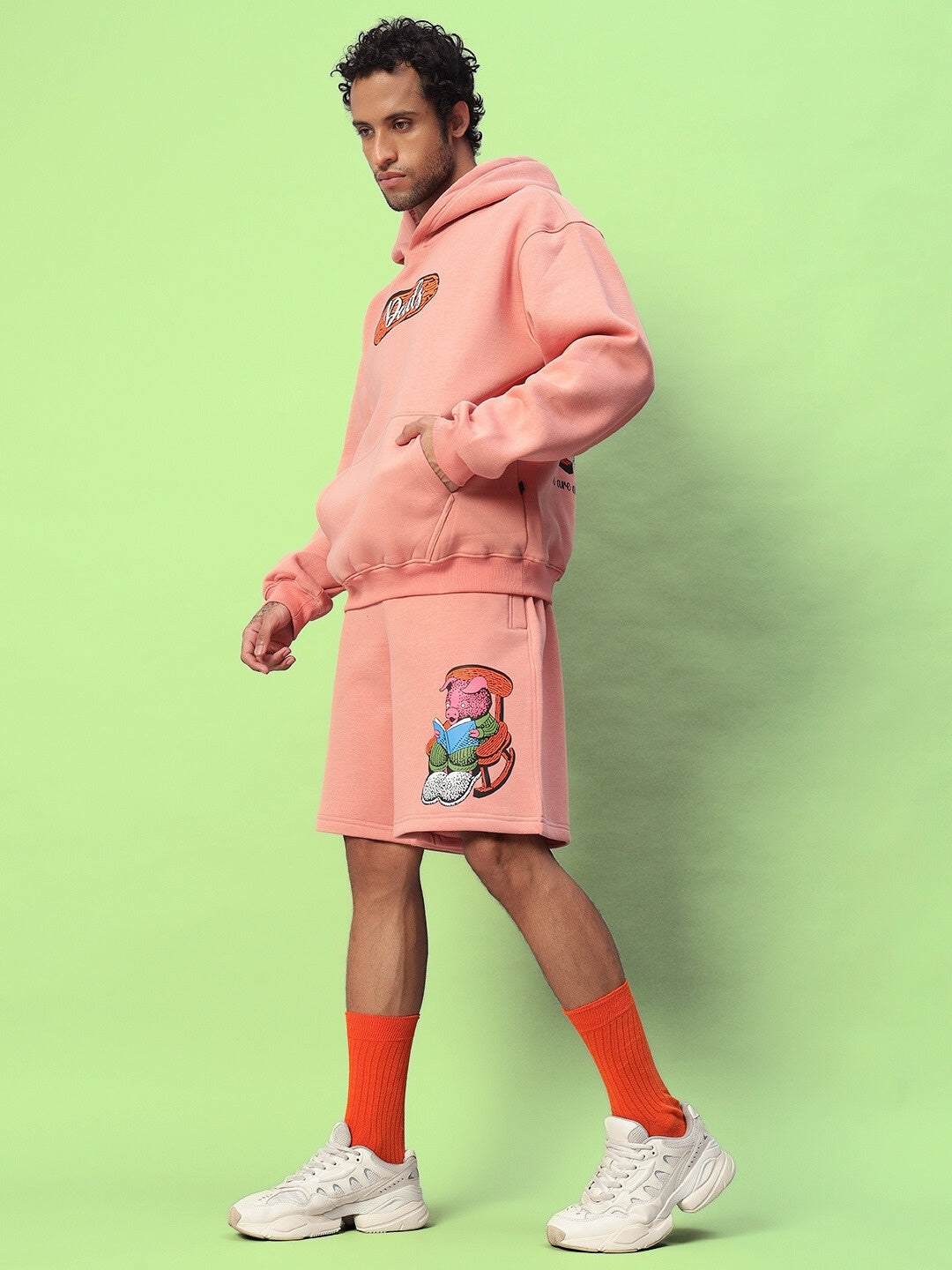 Man wearing Deliberate Fleece Hoodie in Peach with matching shorts, standing in front of a green background.