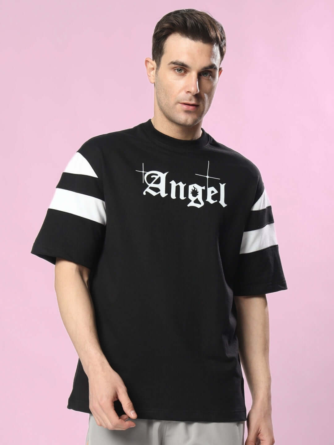 Man wearing DIVINE SPIRIT OVERSIZED T-SHIRT BLACK with 'Angel' text on chest and white striped sleeves, against a pink background.