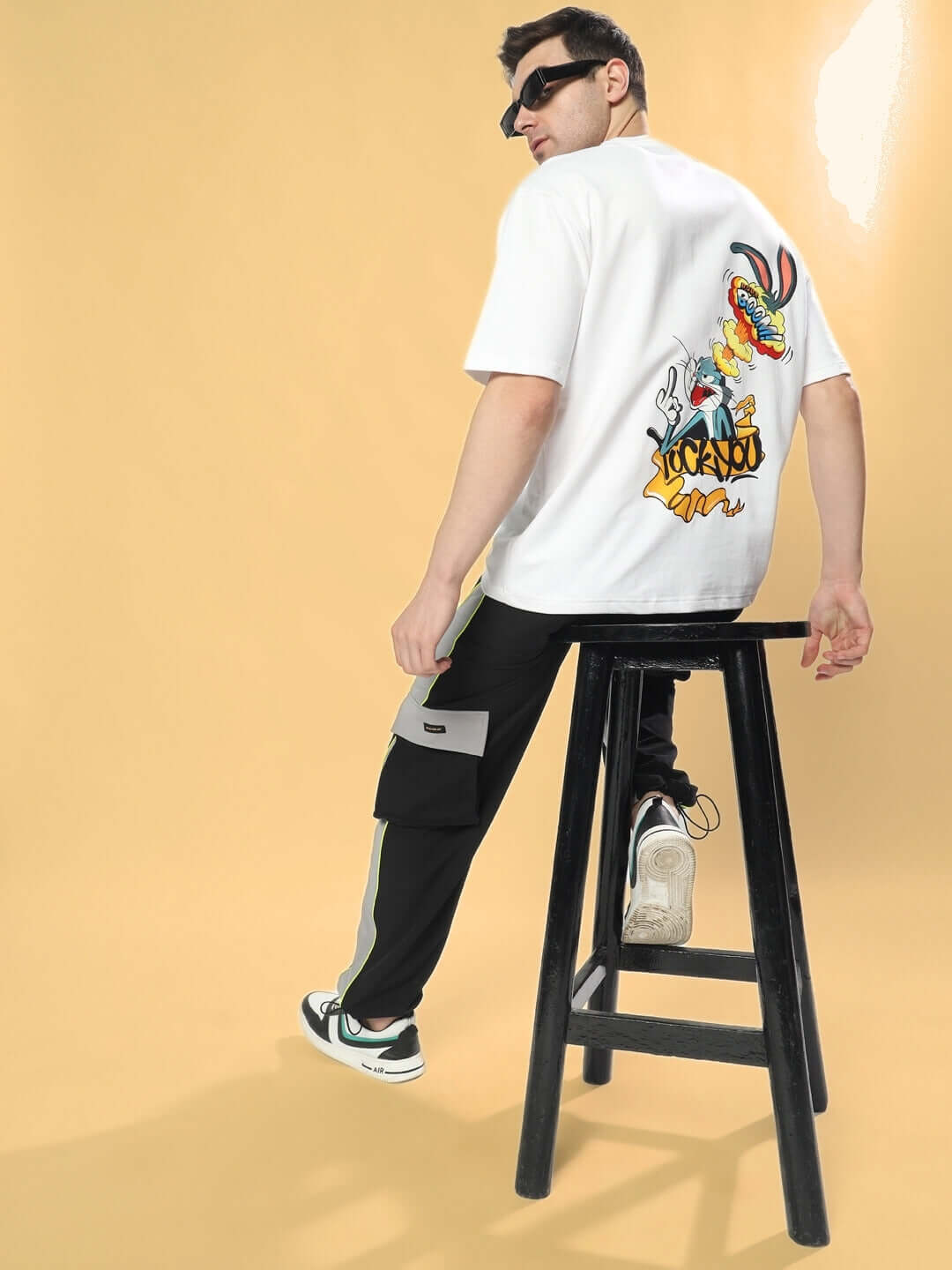 Model wearing BOOM BUNNY OVER-SIZED T-SHIRT WHITE with cartoon design, seated on a black stool.