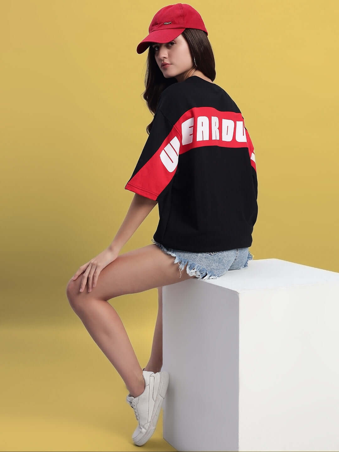 Model wearing Women's Titan Over-Sized T-Shirt in black-red, seated on a white cube, paired with denim shorts, white sneakers, and red cap.