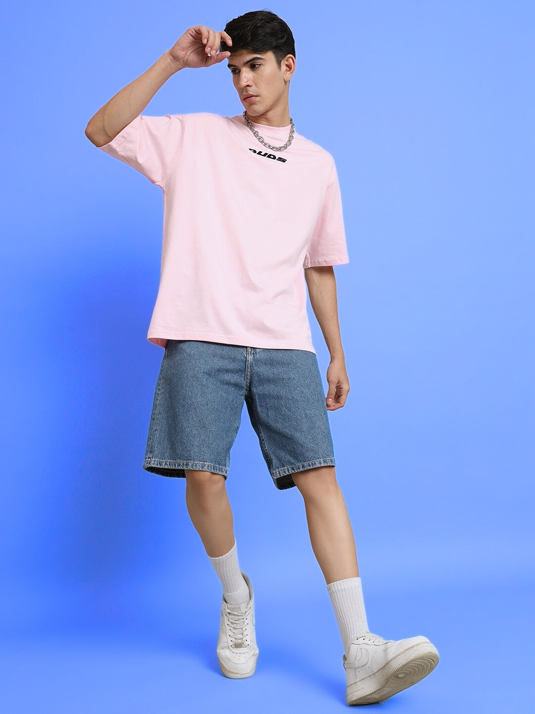 Man wearing a TOP-DOG oversized pink t-shirt with denim shorts and white sneakers, standing against a blue background.