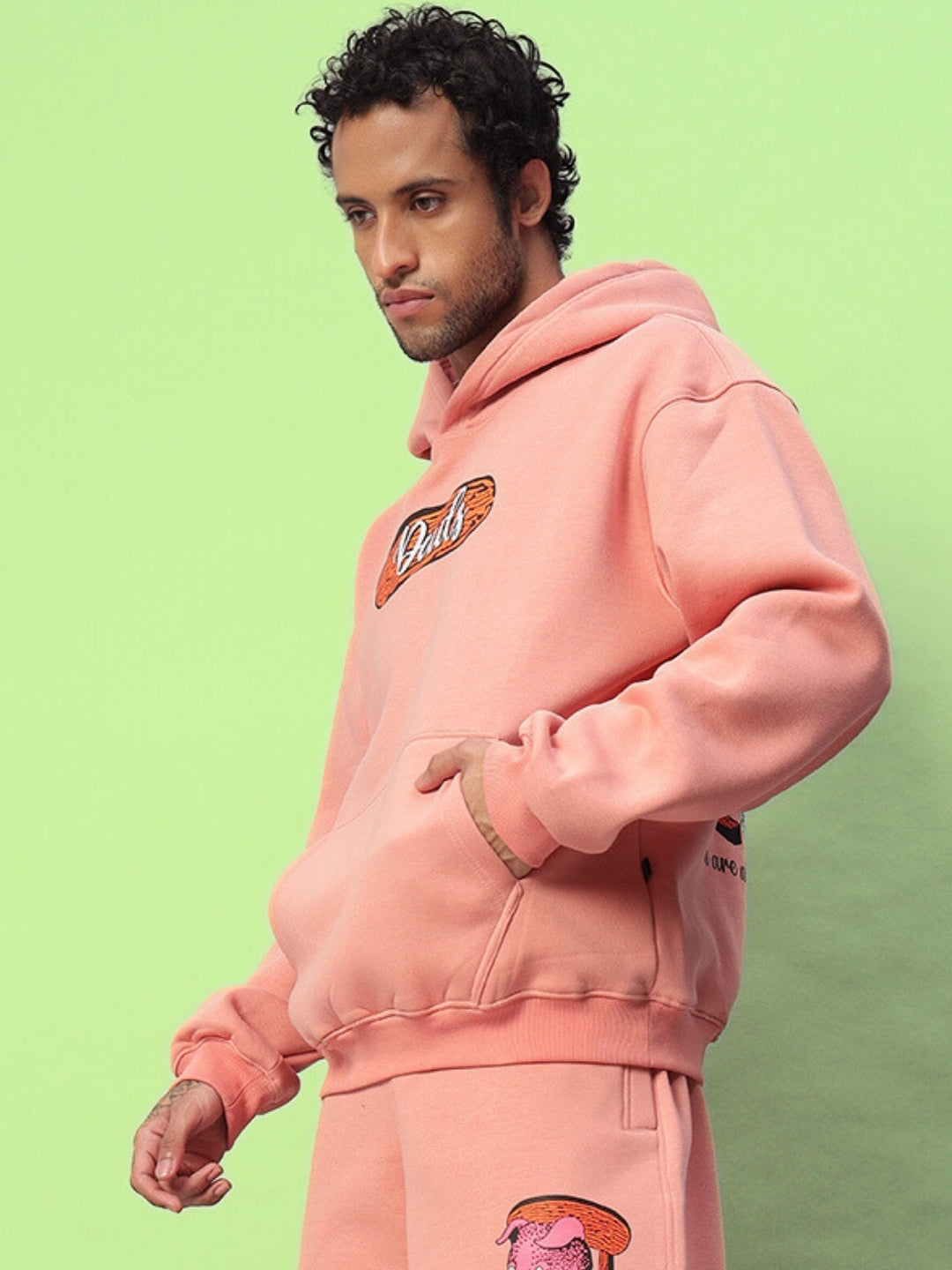 Man wearing a Deliberate Fleece Hoodie in peach color, featuring stylish design and logo, standing against a light green background.
