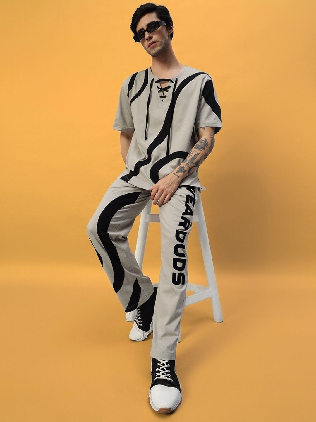 Model wearing grey oversized t-shirt with black surfing design, paired with matching pants, sitting on a white stool against an orange background.