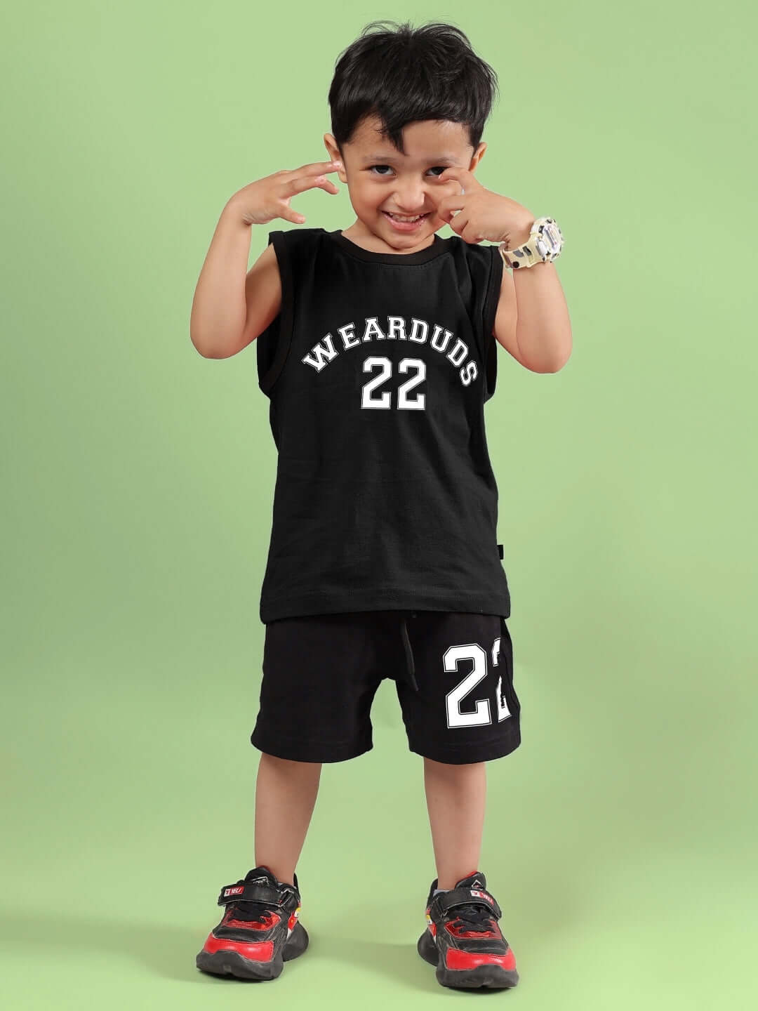 Young boy wearing a black AMIGA co-ord by DUDS for boys and girls, featuring a matching top and shorts with the number 22, smiling on a green backdrop.