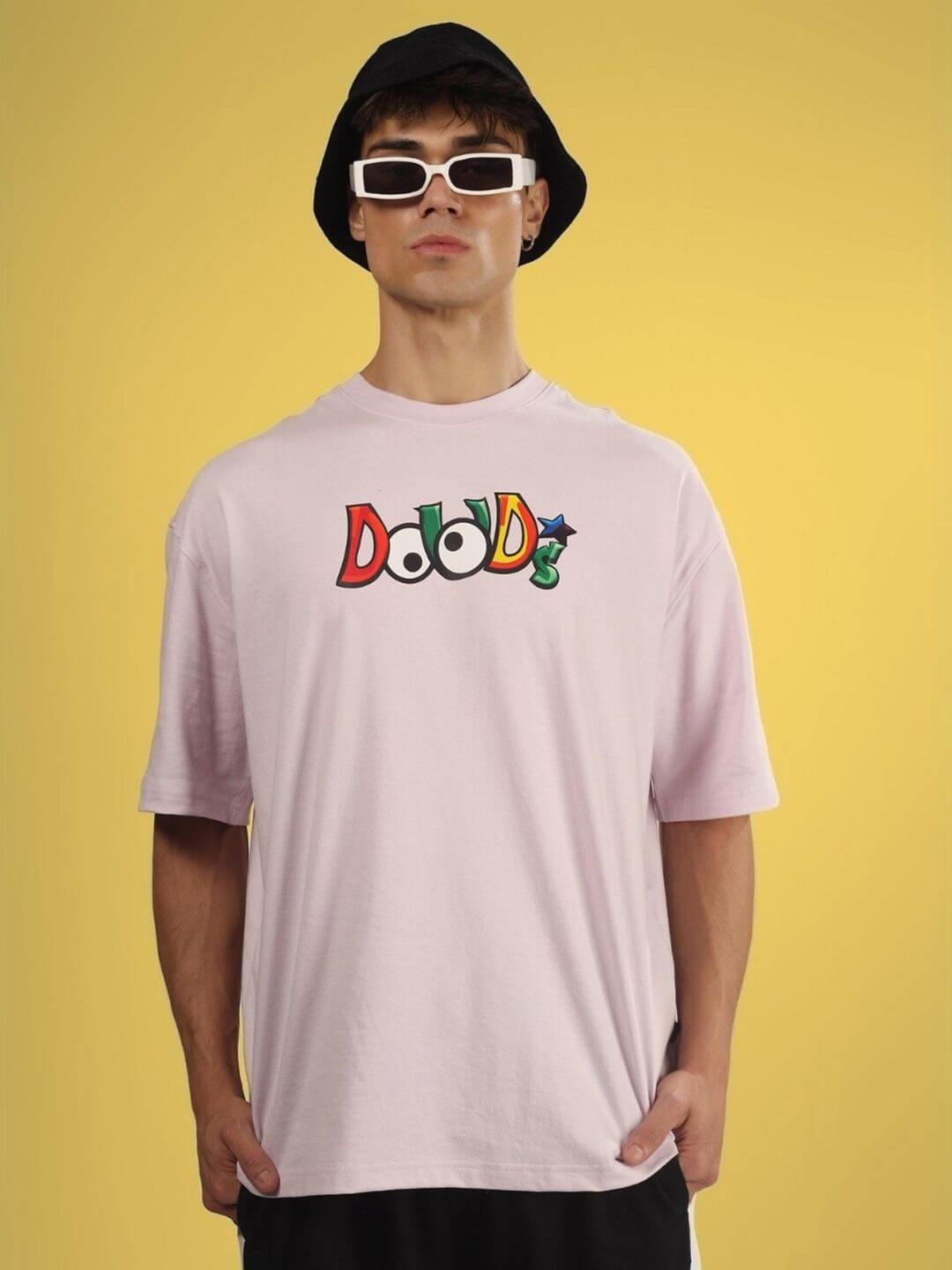 Man wearing DUDS MONSTER OVER-SIZED T-SHIRT PALE PINK with colorful logo, sunglasses, and a hat on yellow background.