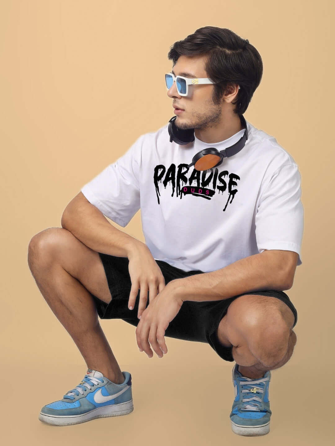 Model wearing Fulness Paradise Summer Shorts in black, paired with a white graphic tee and blue sneakers, sitting on a tan background.
