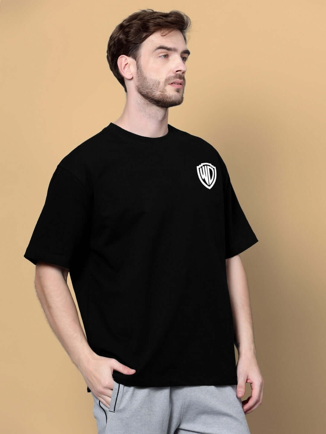 Man wearing POLICIA OVER-SIZED T-SHIRT BLACK with logo on chest, beige background.