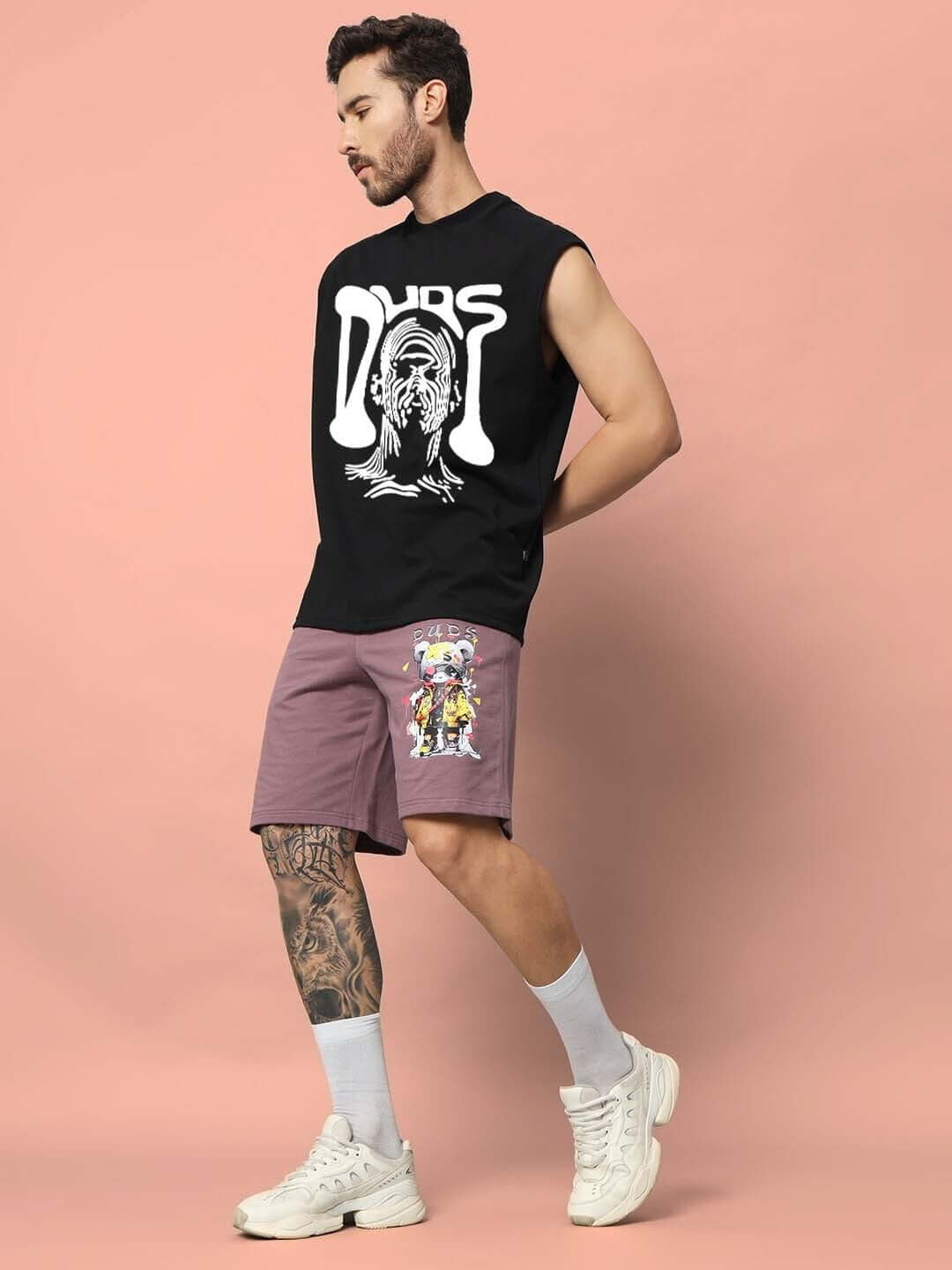 Man wearing DUDS Taska black gym t-shirt, casual shorts, and white sneakers against a pink background.