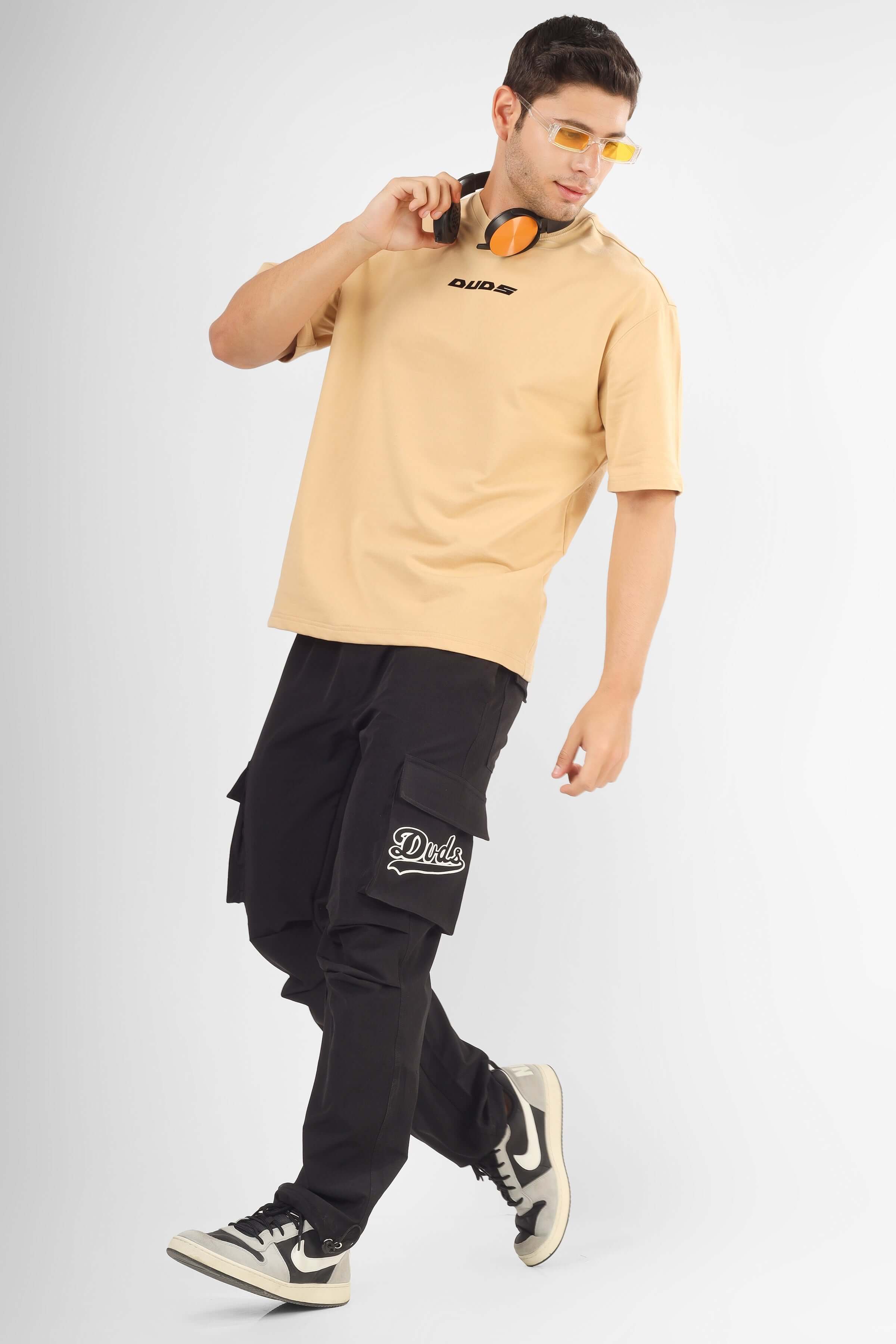Man wearing POSITIVE MINDSET OVER-SIZED T-SHIRT NUDE with cargo pants and sneakers, holding orange headphones.