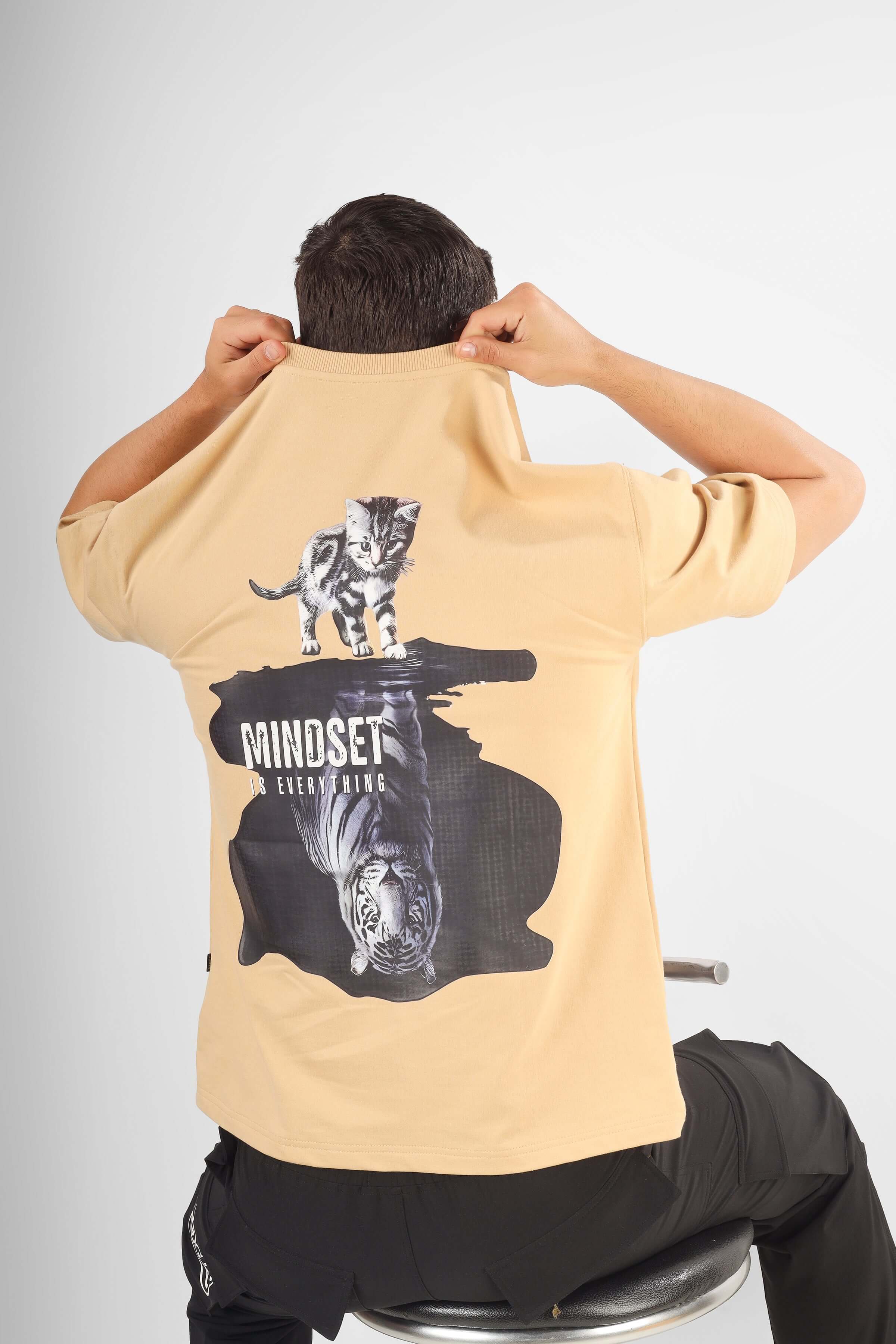 Rear view of person wearing POSITIVE MINDSET OVER-SIZED T-SHIRT NUDE with unique graphic design, sitting on a stool.