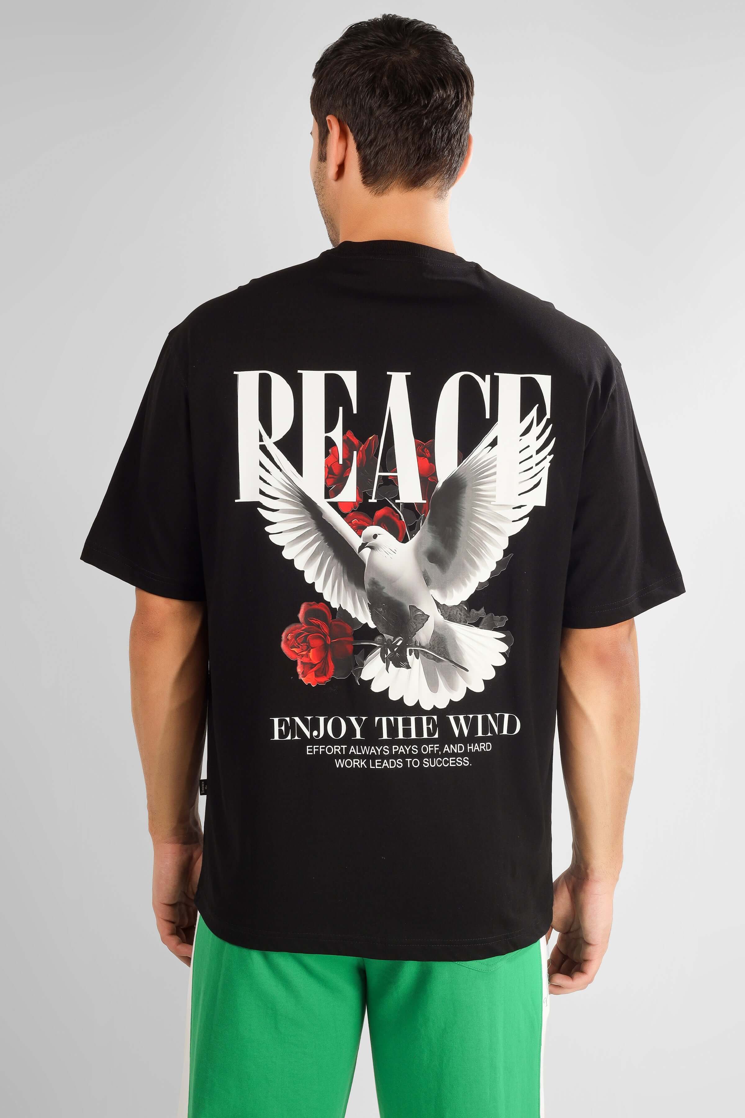 Back view of model wearing ENJOY THE WIND OVER-SIZED T-SHIRT BLACK with dove and peace print on the back.