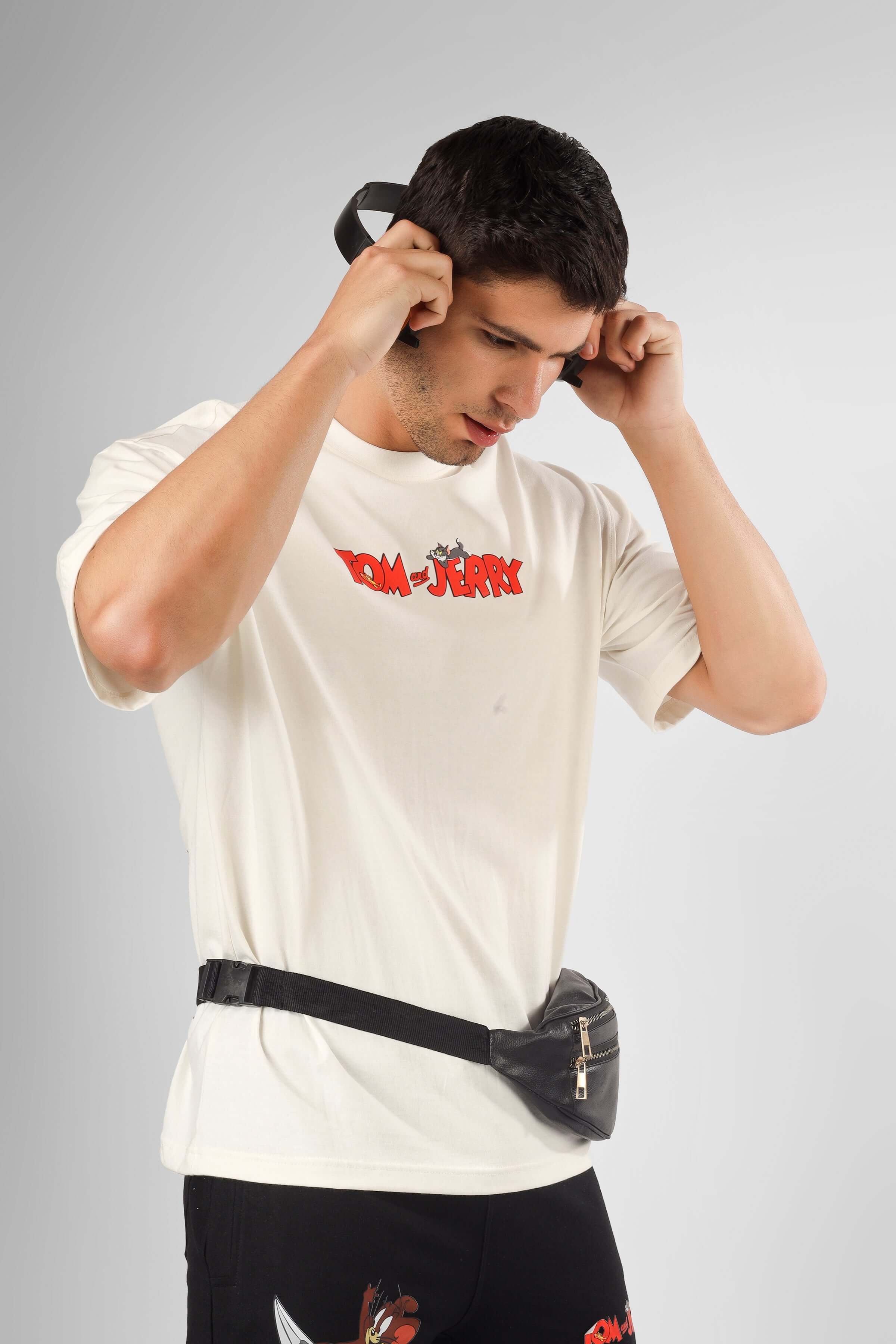 Man wearing TOM & JERRY OVER-SIZED T-SHIRT CREAM with black fanny pack and adjusting headphones