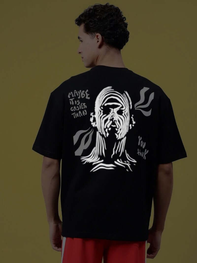 Man wearing MAYBE IT IS EASIER THAN YOU THINK REFLECTOR T-SHIRT BLACK with reflective text design, standing against yellow background.