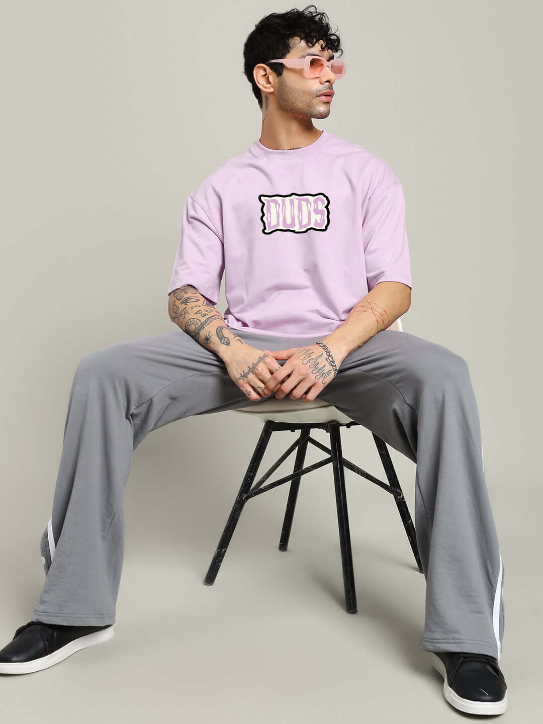 Man wearing a lavender oversized t-shirt with printed text 'Let's Get Spooky', paired with gray pants and pink tinted sunglasses, seated on a chair.