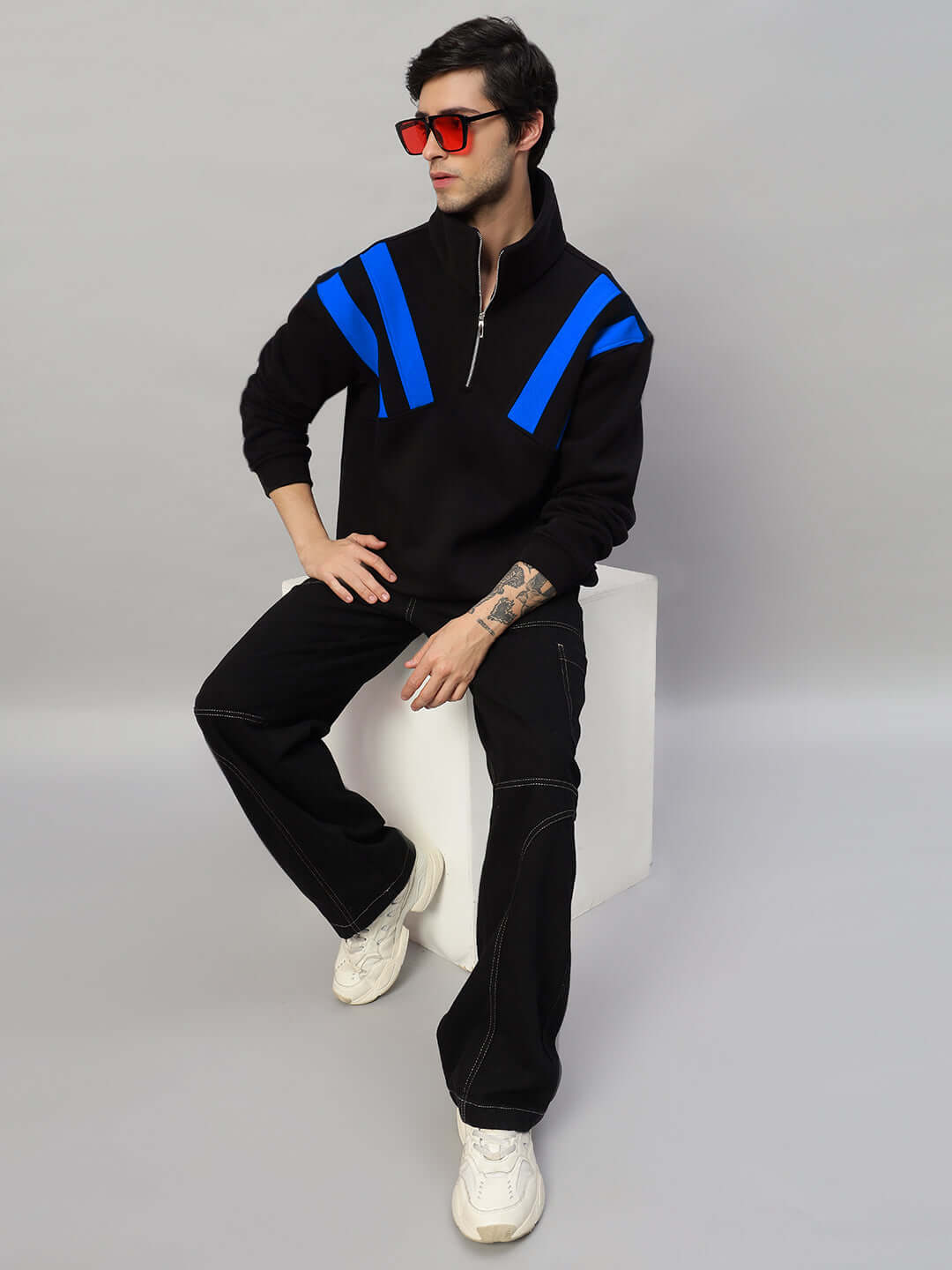 Model wearing CROSSBAR FLEECE COLORBLOCK JACKET in black and blue, showcasing modern design and luxurious comfort, sitting on a cube.