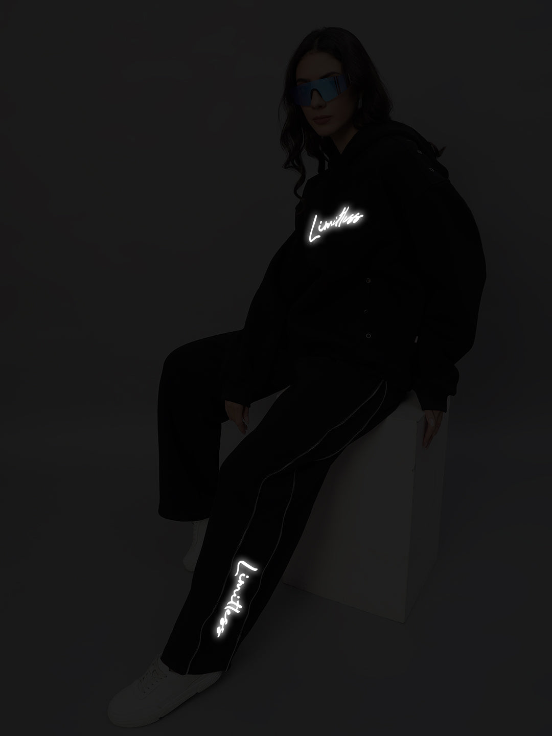 Model wearing Women's Limitless Fleece Reflector Co-Ord in Black with reflective details, sitting on a white block against a dark background.