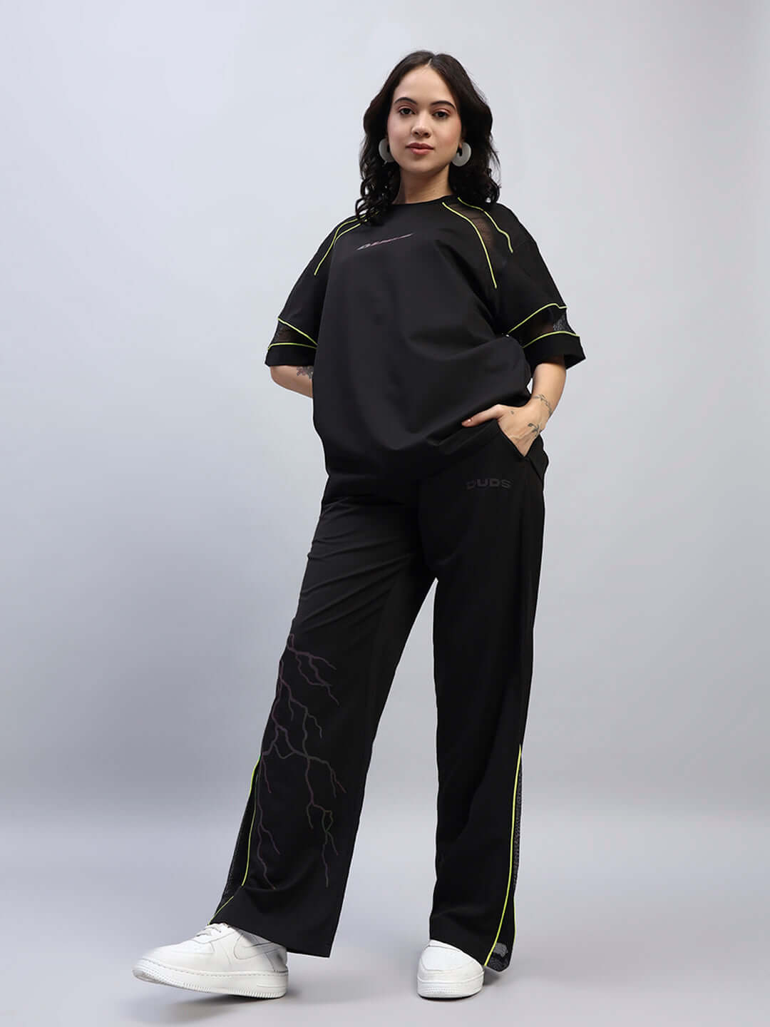 Woman wearing black relaxed-fit joggers and matching top with neon accents, standing confidently against a neutral background.
