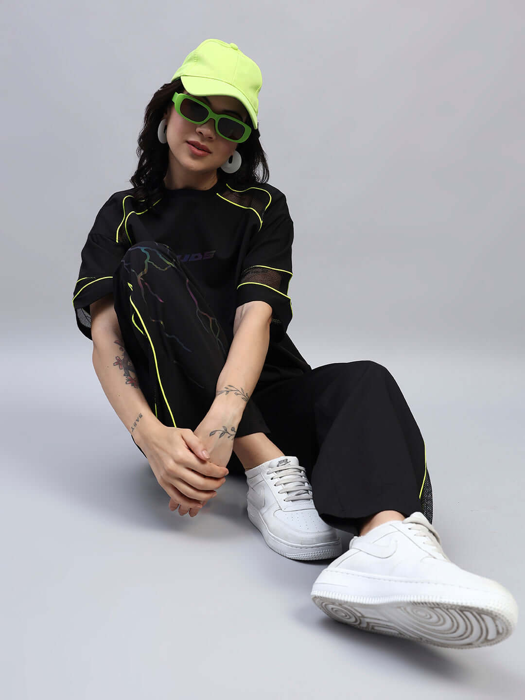 Model wearing black relaxed-fit joggers with neon accents, matching t-shirt, lime green cap and sunglasses, white sneakers.