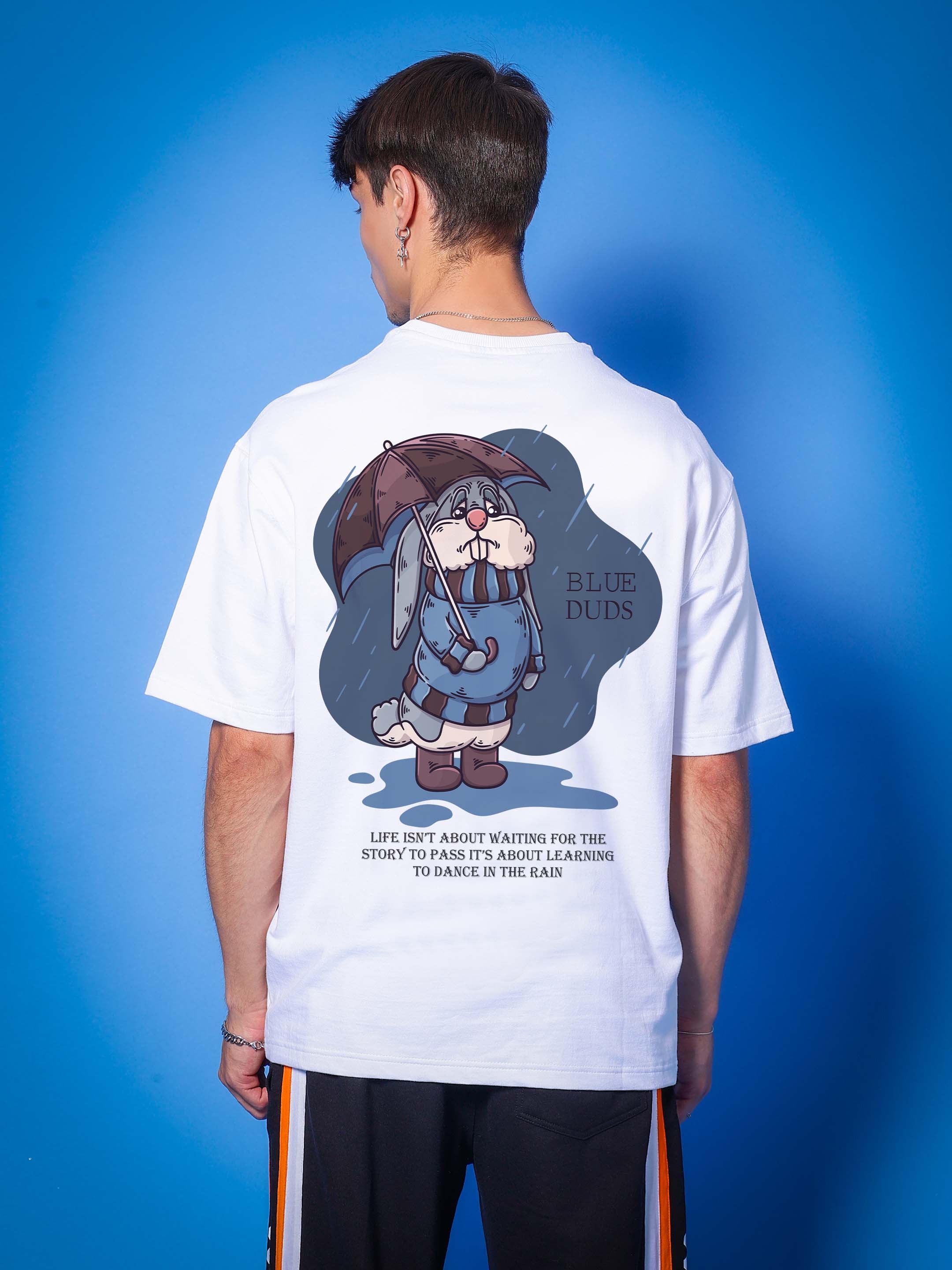 Man wearing BLUE LAPIN OVER-SIZED T-SHIRT WHITE with cartoon graphic and inspirational quote on the back, blue background.