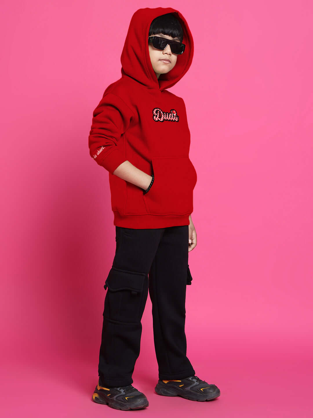 Child wearing a stylish red cupcake co-ord outfit with hoodie and black cargo pants against a pink background.