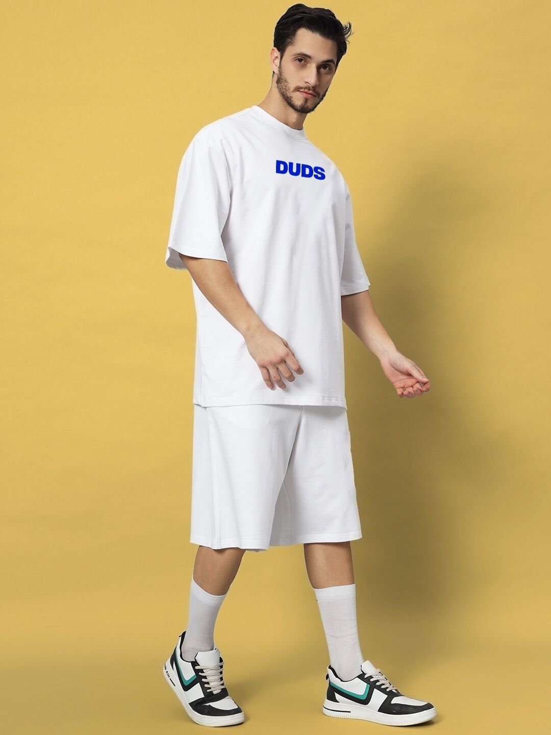 Man wearing ABSENT PRESENT CO-ORD SET WHITE t-shirt and shorts, showcasing modern minimalism and style against a yellow background.