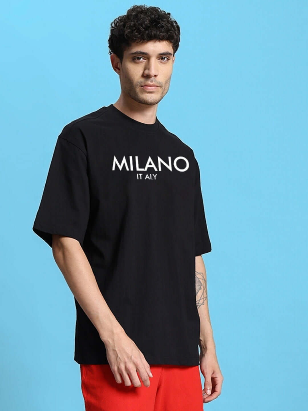 Man wearing a black oversized t-shirt with 'MILANO ITALY' print, styled with red pants against a blue background.