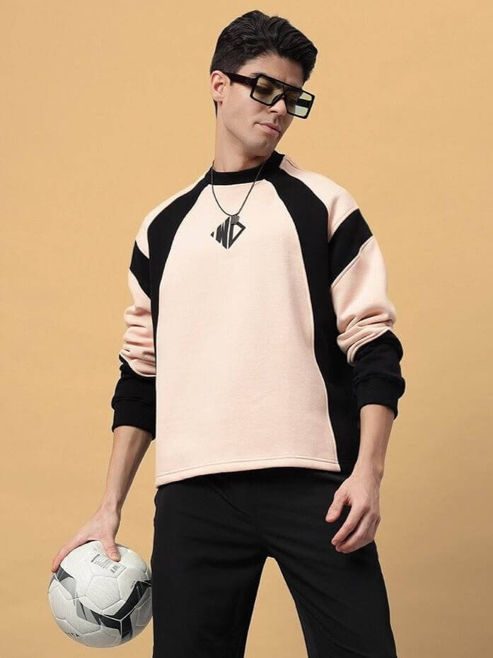 Man wearing beige and black sweatshirt holding a soccer ball, looking stylish and casual.