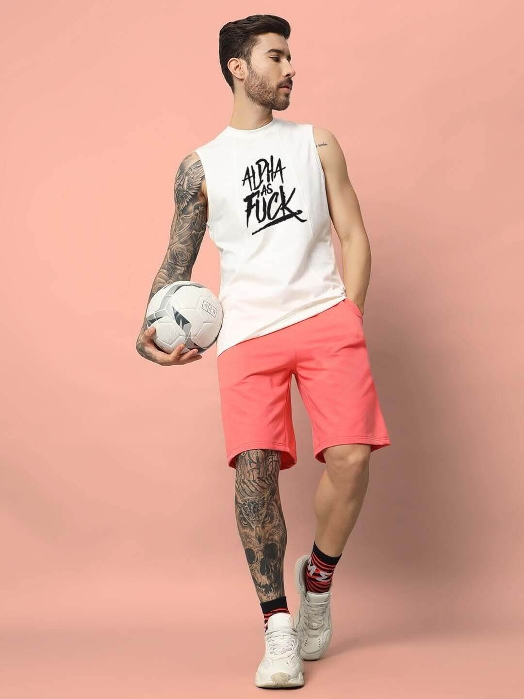Man wearing an off-white 'ALPHA AS FUCK' gym t-shirt holding a soccer ball with a stylish tattoo sleeve, standing against a pink background.