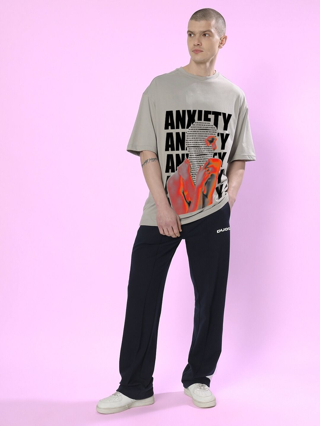 Model wearing ANXIETY OVER-SIZED T-SHIRT GREY with graphic print on front, styled with black pants, against a pink background.