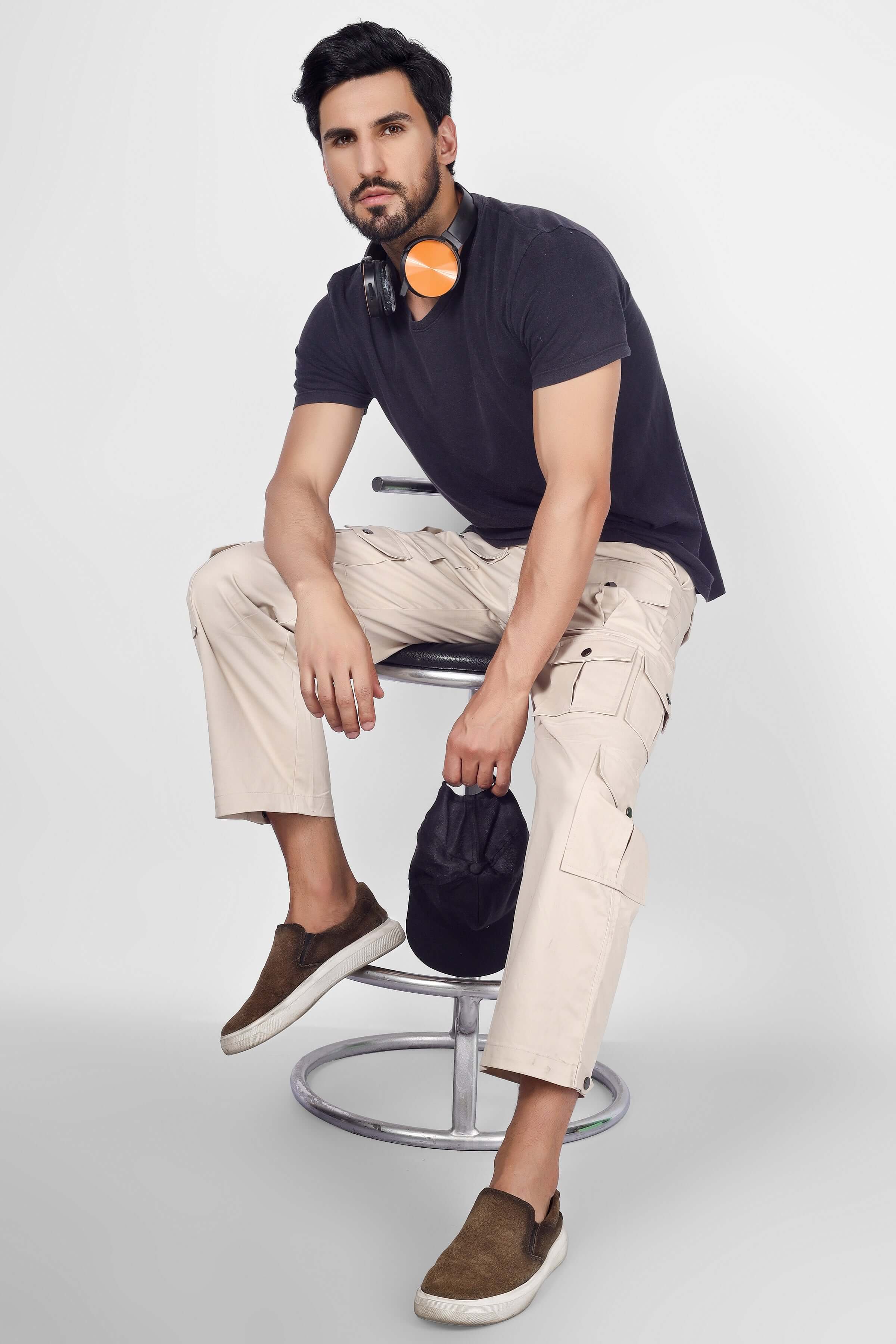 Man wearing Arcadia White 14 Pocket Cargo Pants, sitting on a stool with headphones around neck, showcasing casual style. Fashionable and practical.