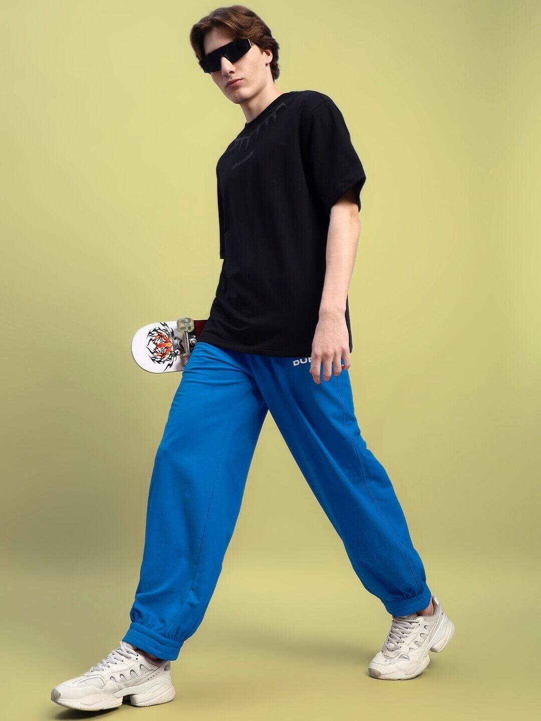 Man wearing AUSTRIAN RELAXED-FIT JOGGERS BLUE holding a skateboard, showcasing a stylish athleisure outfit on a light background.