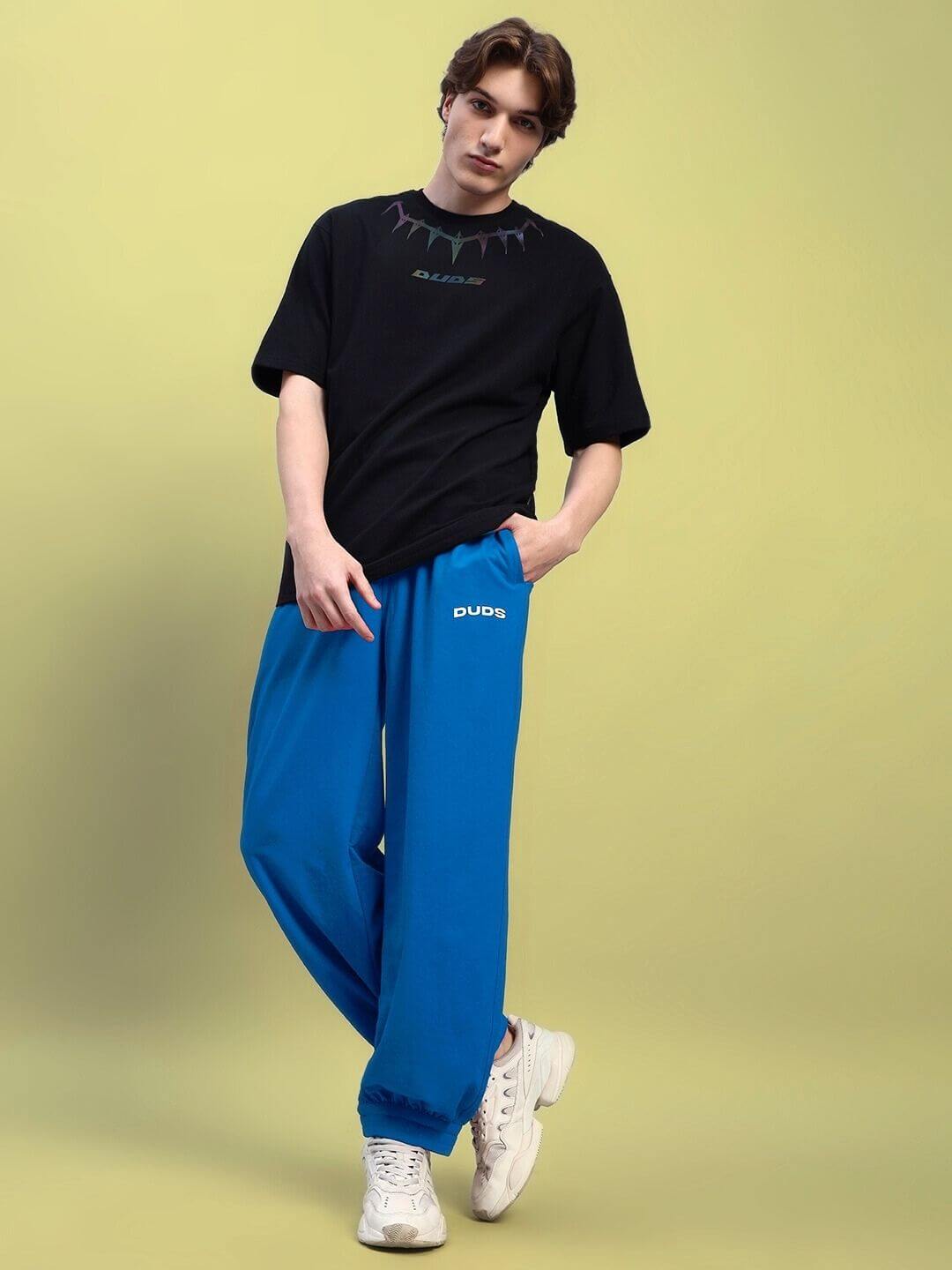 Model wearing AUSTRIAN RELAXED-FIT JOGGERS BLUE with white sneakers on a yellow background, featuring an elastic waistband and drawstrings.