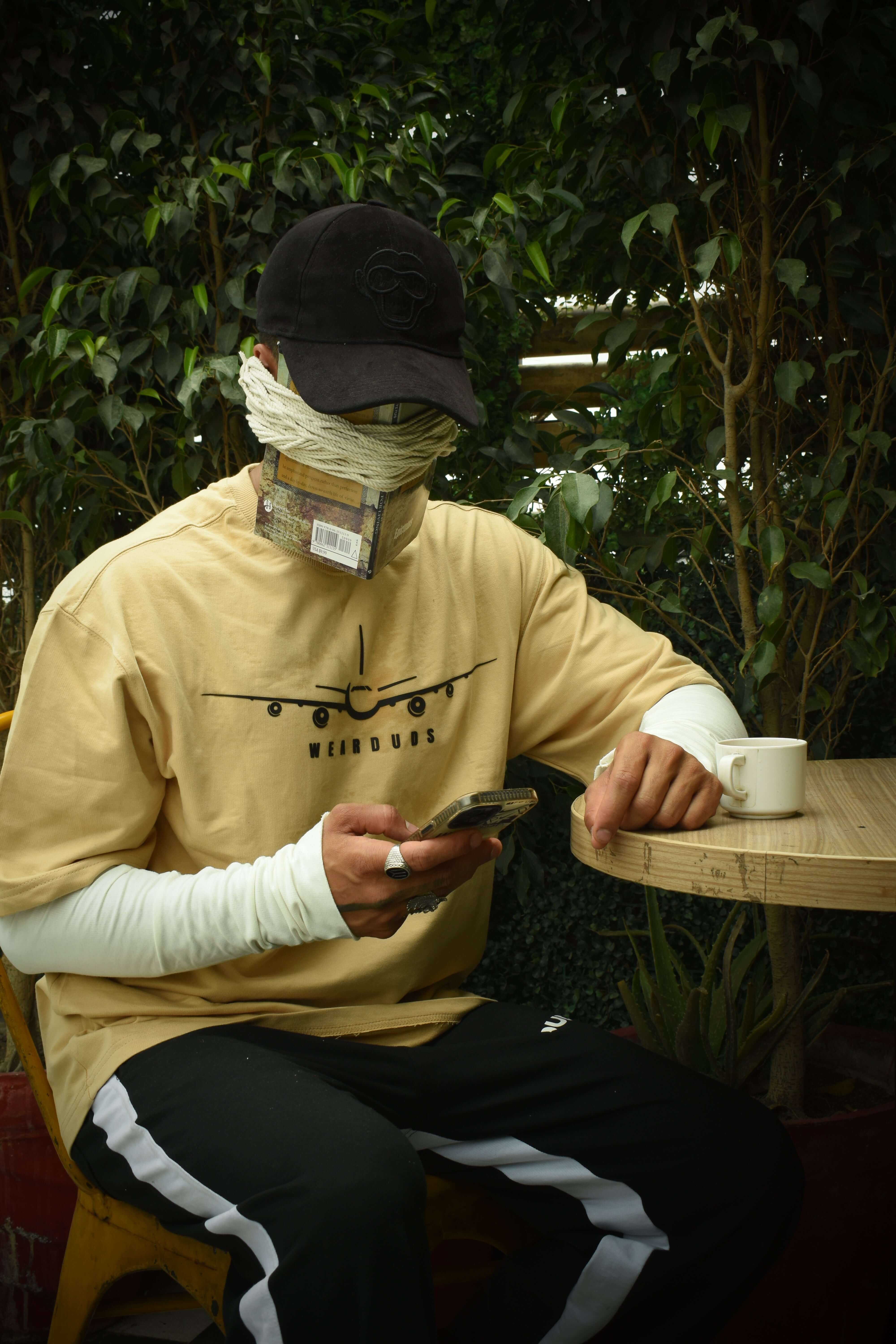 Person wearing a beige aviator designer oversized T-shirt with graphic print, sitting at outdoor cafe table with coffee, checking phone.