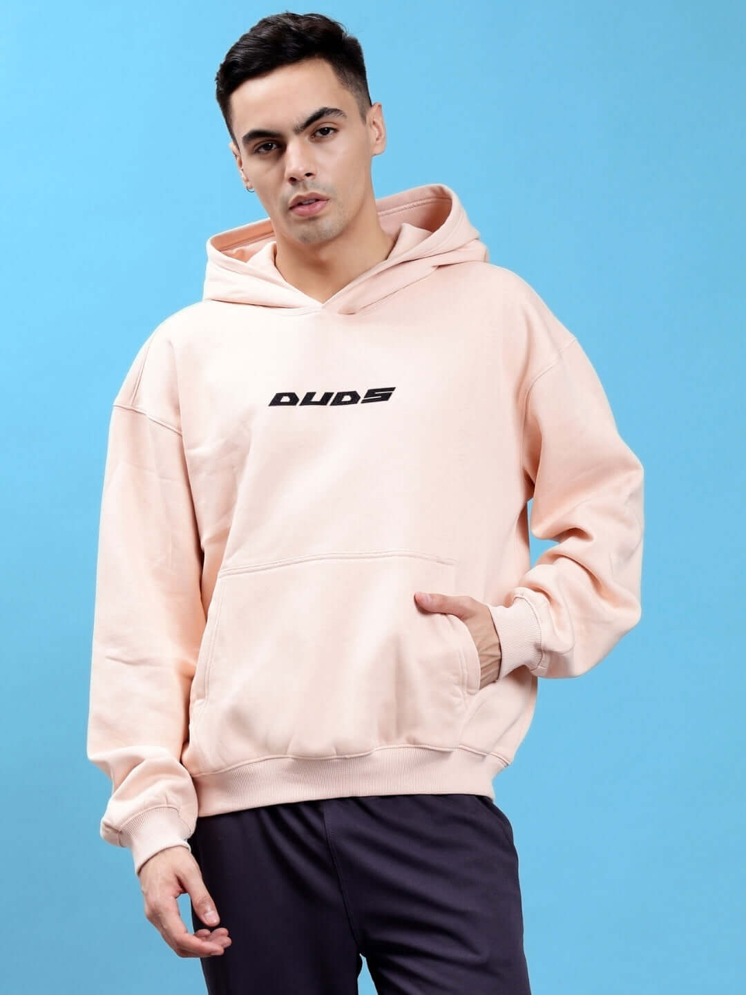 Model wearing HUG OVER-SIZED COTTON FLEECE HOODIES BEIGE with soft style logo, showcasing streetwear fashion against blue background.