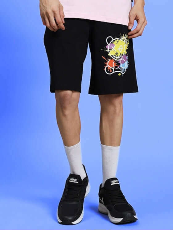 Person wearing BE KIND REGULAR FIT SHORTS BLACK with graphic design, showcasing modern casual style, against blue background.