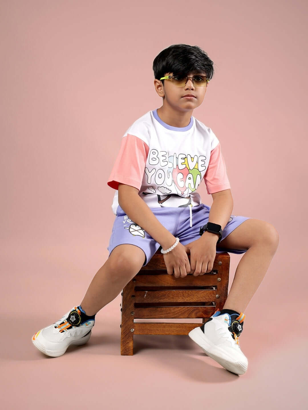 Boy wearing Believe T-shirt in white-pink, seated on a wooden stool against a pink background, styled with sunglasses and sneakers.