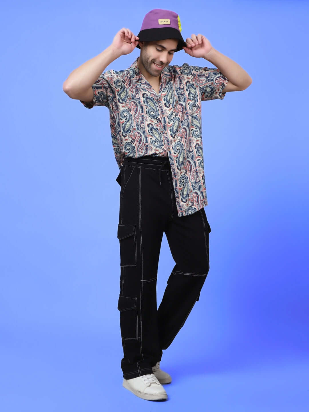 Model wearing black carpenter cargo pants and paisley shirt with purple hat, showcasing casual fashion style against blue background.
