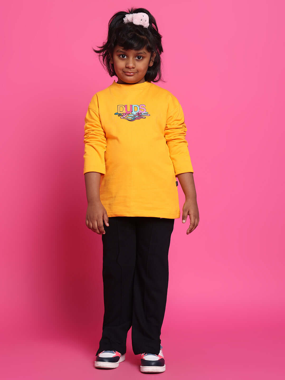 Young girl wearing CUBE JOGGER for Boys & Girls in black by DUDS, standing against a pink background, showcasing comfortable fashion.