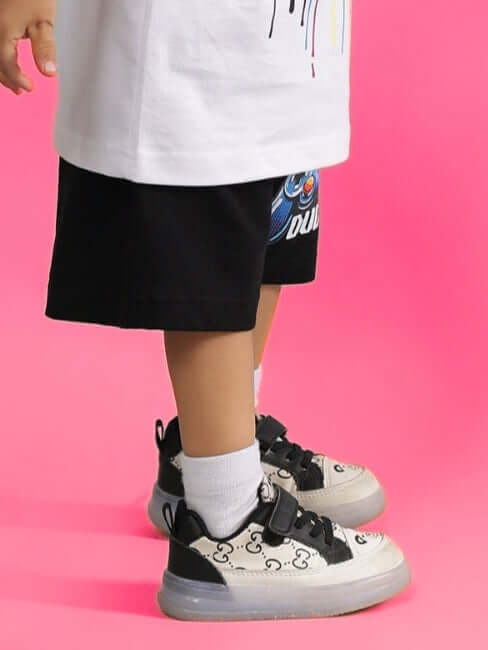 Child wearing GAME TIME SHORTS FOR BOYS & GIRLS BLACK with printed sneakers against pink background