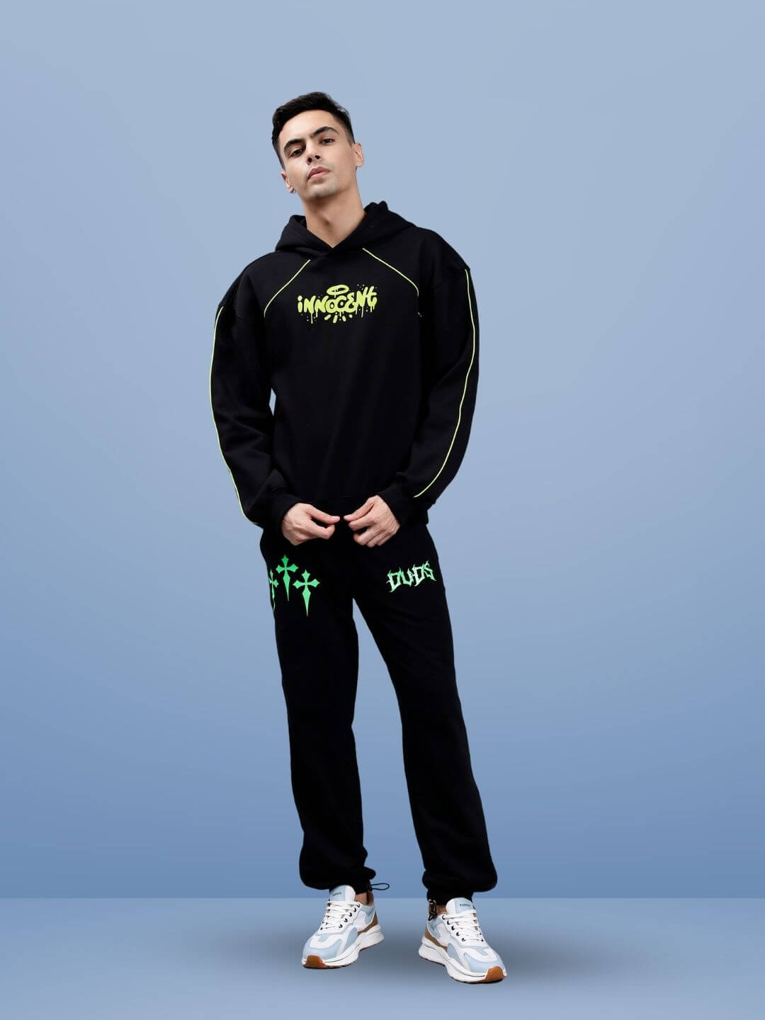 Person wearing black hoodie and joggers set with neon green graphics, standing against a blue background.