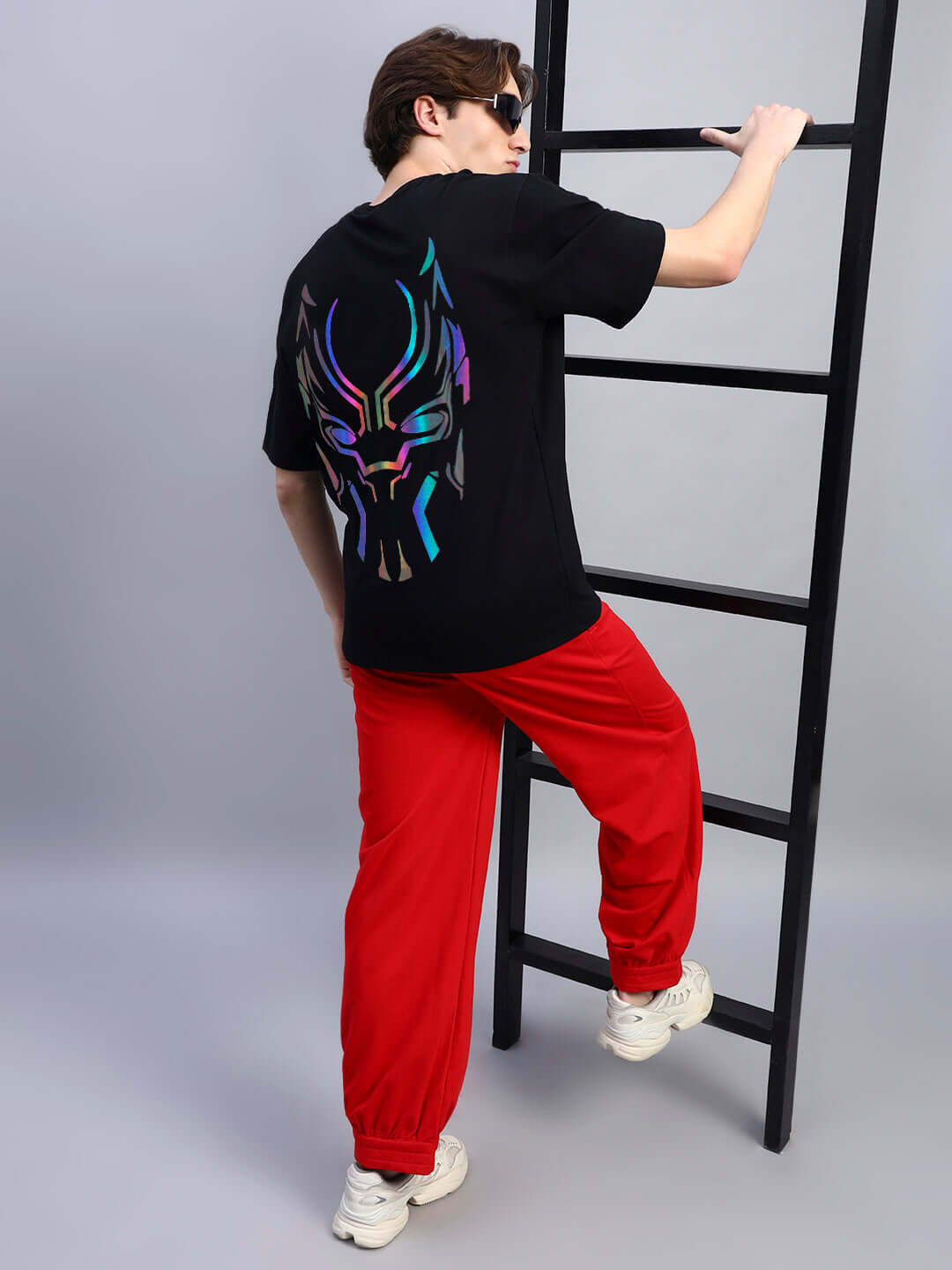 Man wearing Black Panther reflector oversized t-shirt with vibrant back design, paired with red pants, climbing a ladder in studio.