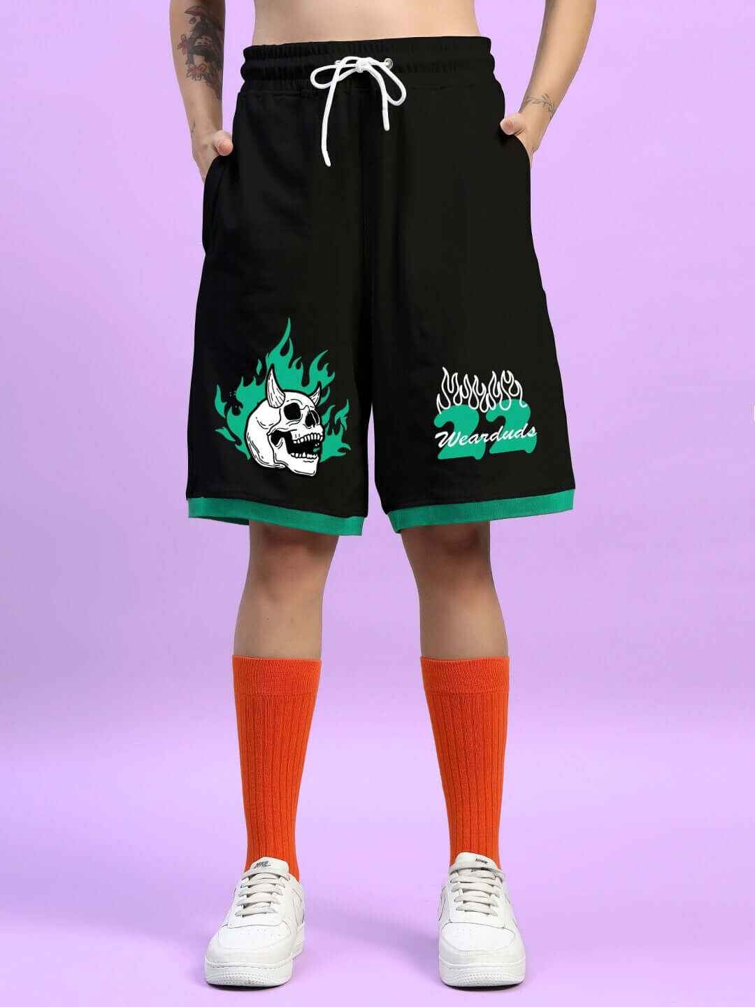 Person wearing black shorts with flame skull print, green trim, and orange socks, standing on light purple background.