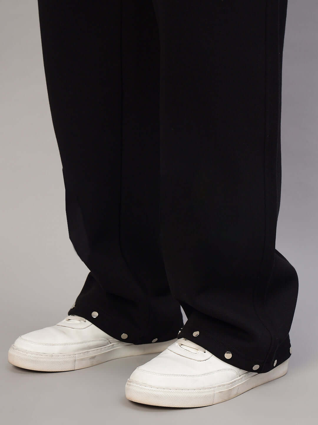 Person wearing black rigid cotton fleece jogger pants with white sneakers, featuring button details at the ankles.