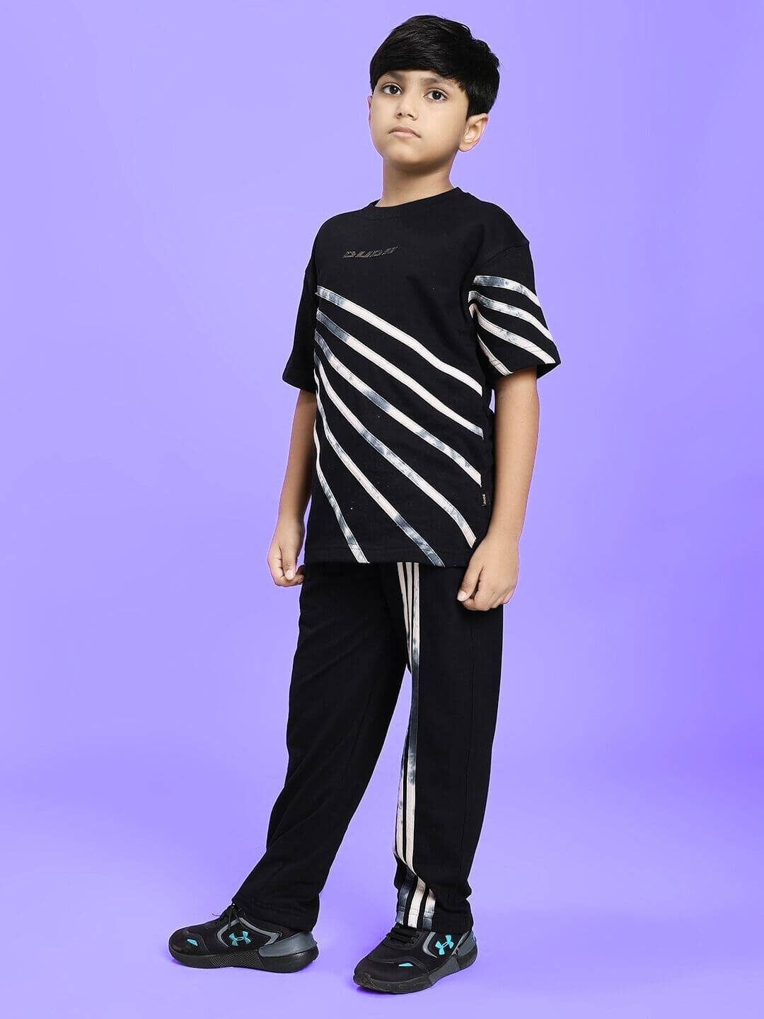 Child wearing FLICKER JOGGER FOR BOYS AND GIRLS BLACK with striped design on pants and top, standing on purple background.
