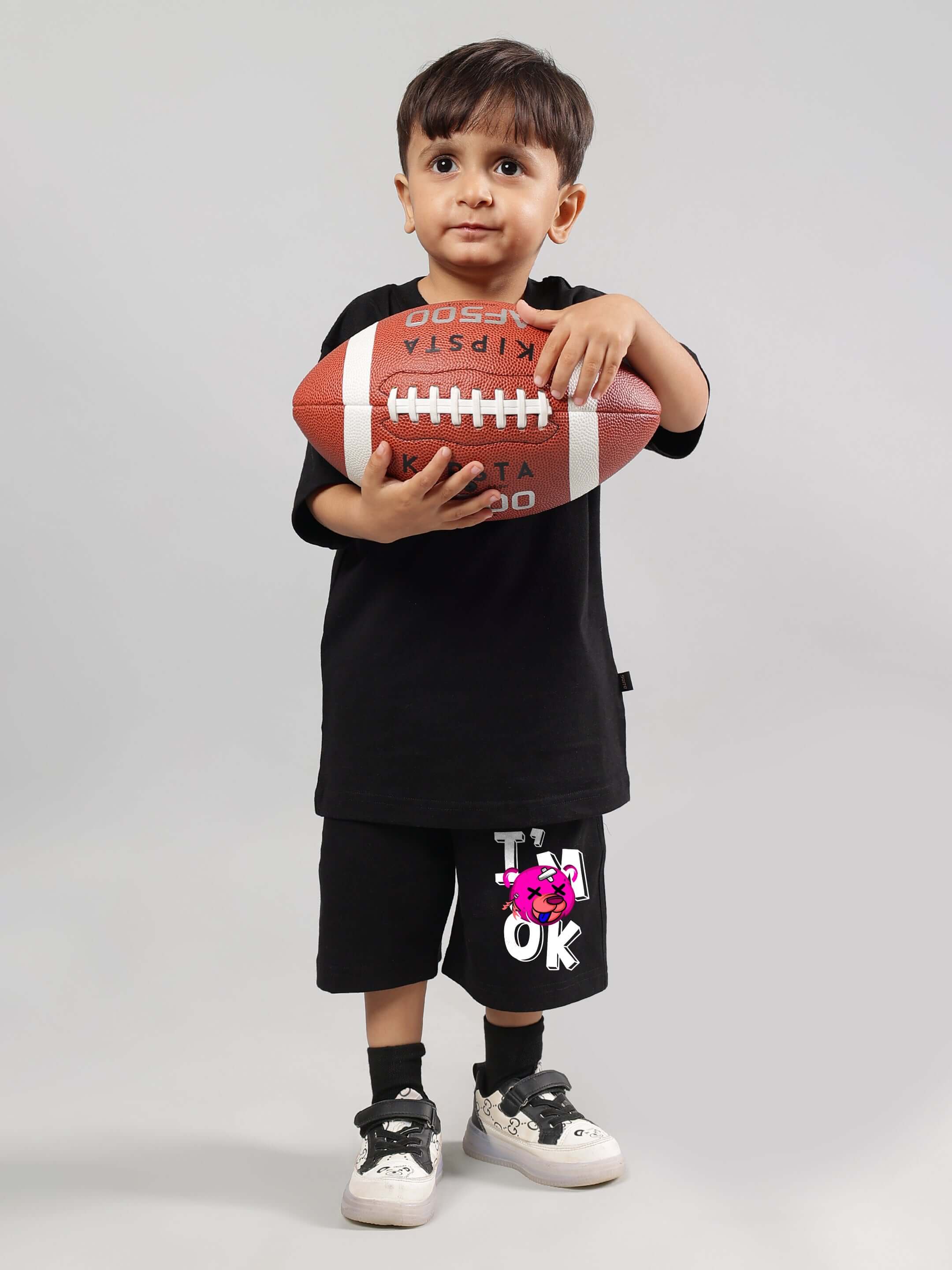 Child wearing black I'M OK T-shirt holding a football, showcasing kids' casual sports apparel.