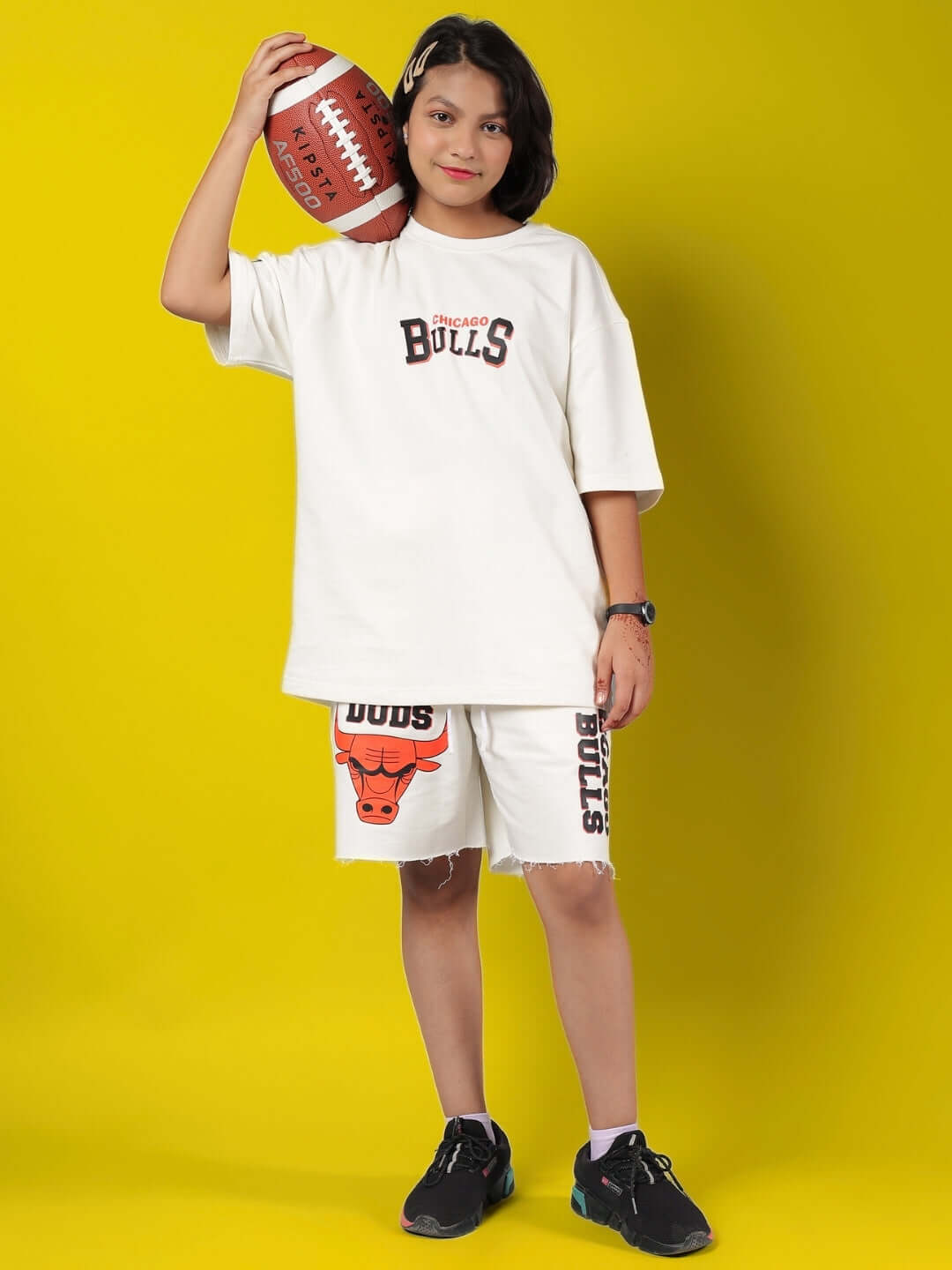 Child wearing BLAZE BULLS SHORTS FOR BOYS & GIRLS OFF-WHITE, holding a football on a yellow background.