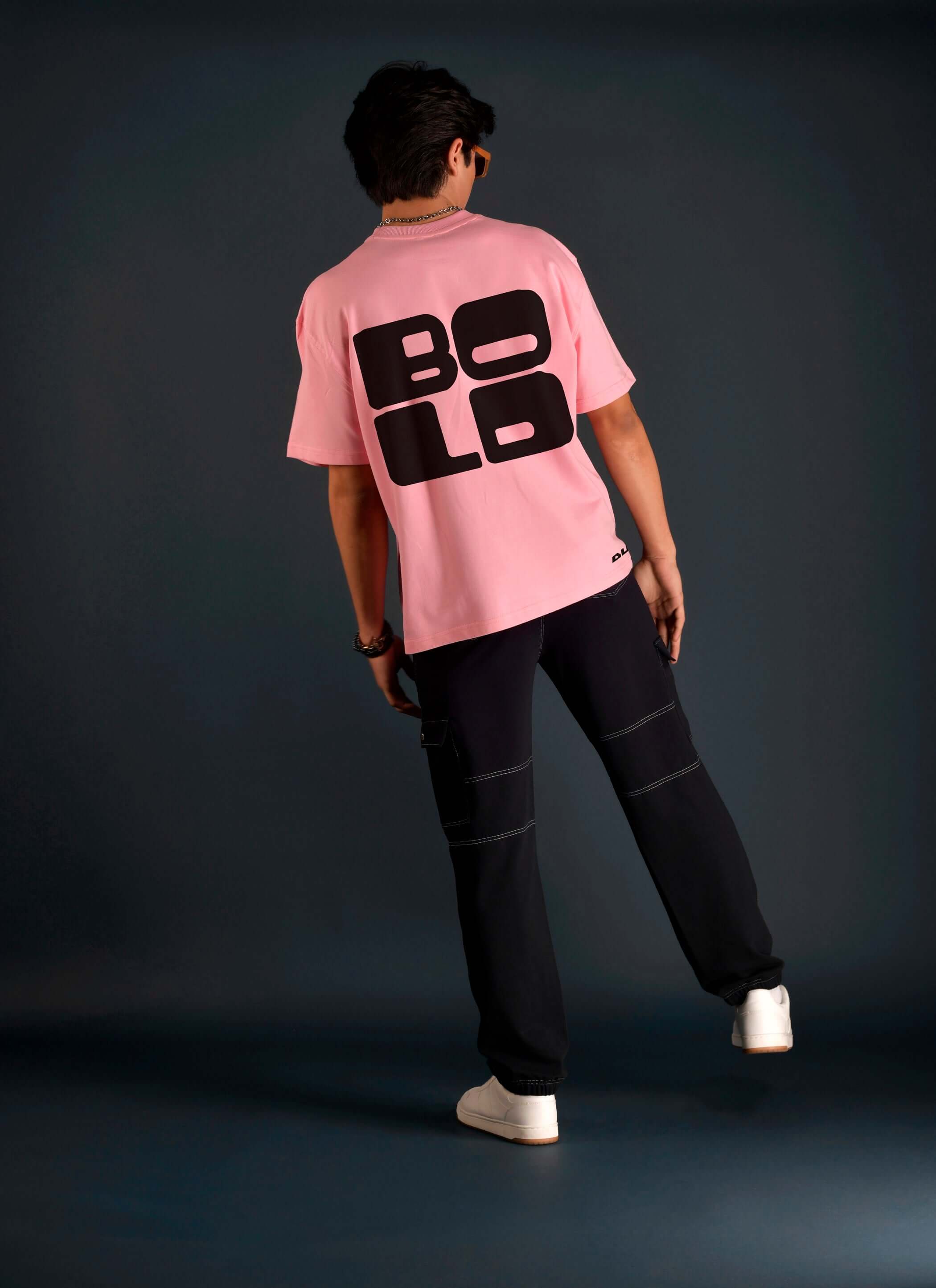 Back view of a person wearing a bold oversized baby pink t-shirt with black lettering, made of premium cotton fabric, paired with dark pants.