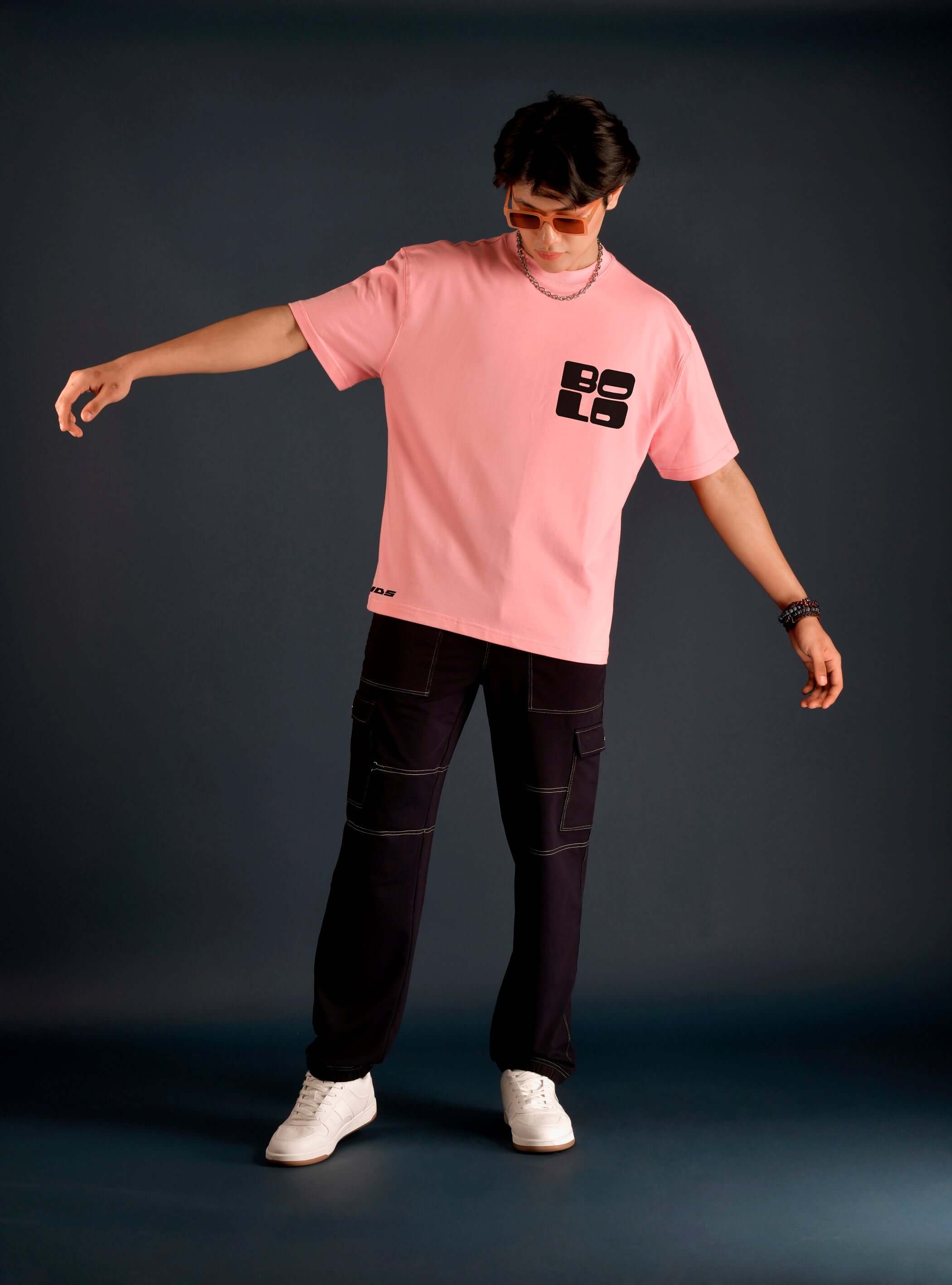 Model wearing a bold oversized baby pink t-shirt with 'BOLD' logo, paired with dark pants and white sneakers, against a dark background.