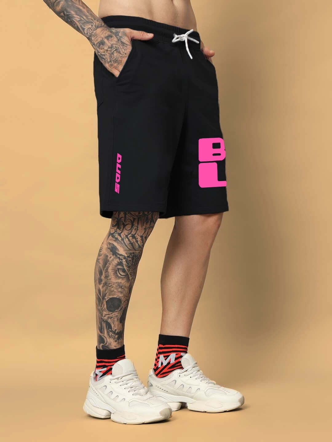 Model wearing black regular fit shorts with bold pink lettering, showcasing tattooed leg in stylish sneakers.