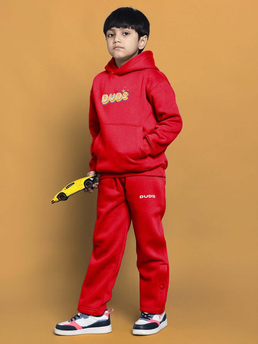 Child wearing a red oversized hoodie with 'Born to Shine' design, holding a toy car, standing against an orange background.